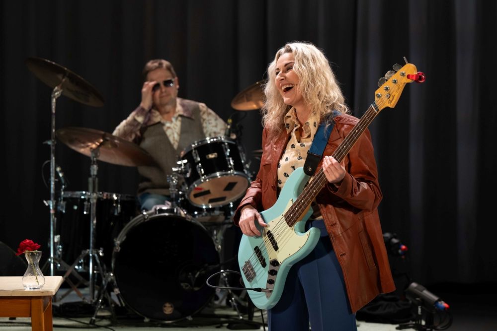 Georgina Field as Sparko smiles, she wears a brown leather jacket and plays a blue guitar
