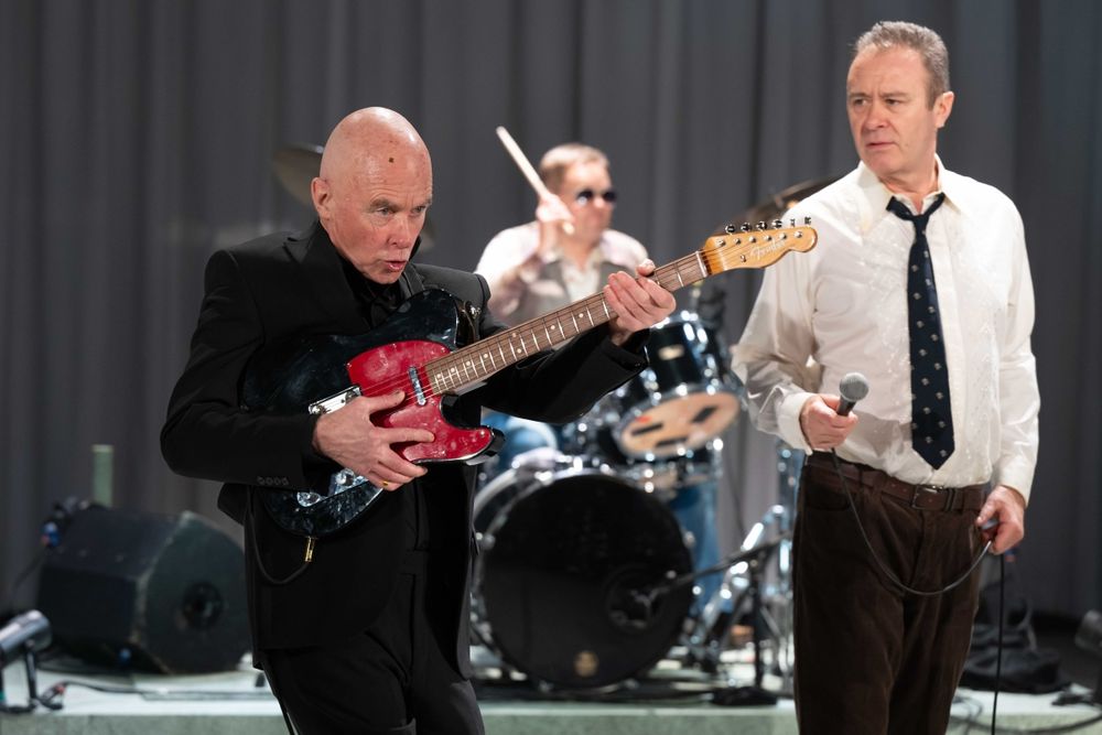 Johnson Willis as Wilko plays a black and red guitar, Jon House as Lee Brilleaux looks on