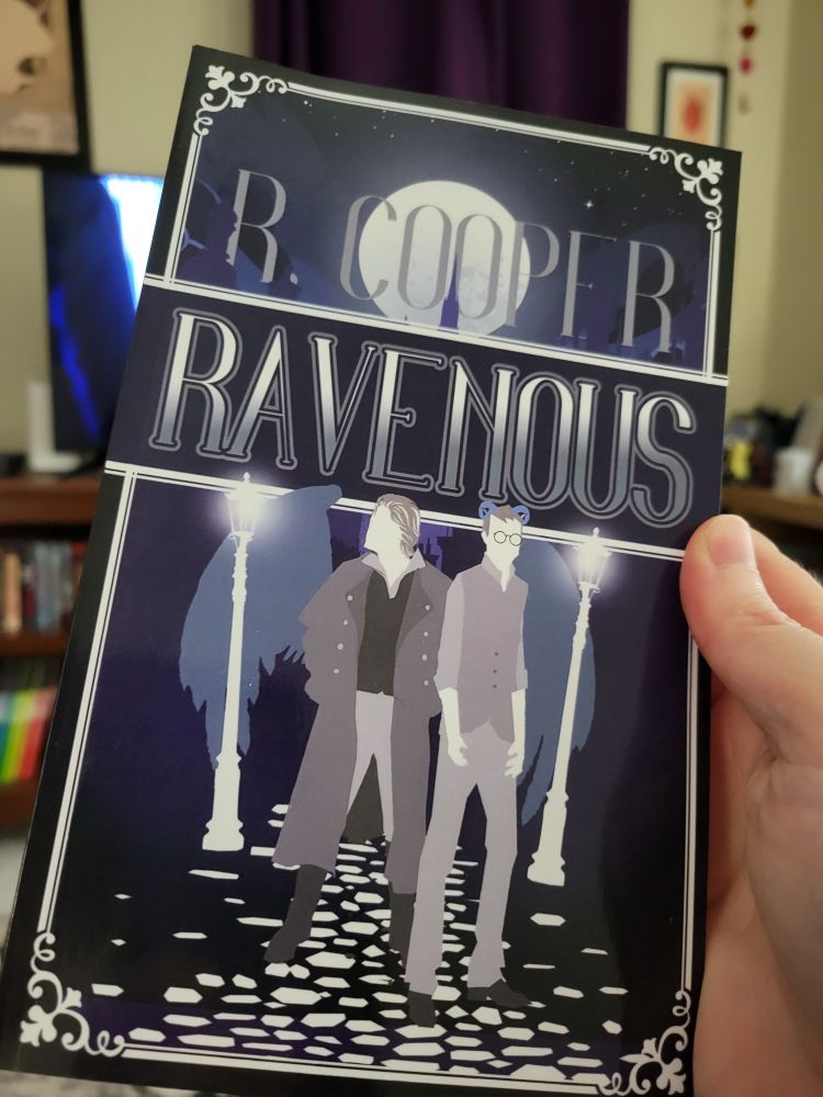 Paperback copy of Ravenous held up in my pale little hand. Cover shows graphics of two men on a moonlit street. One man has horns and the other has wings. 