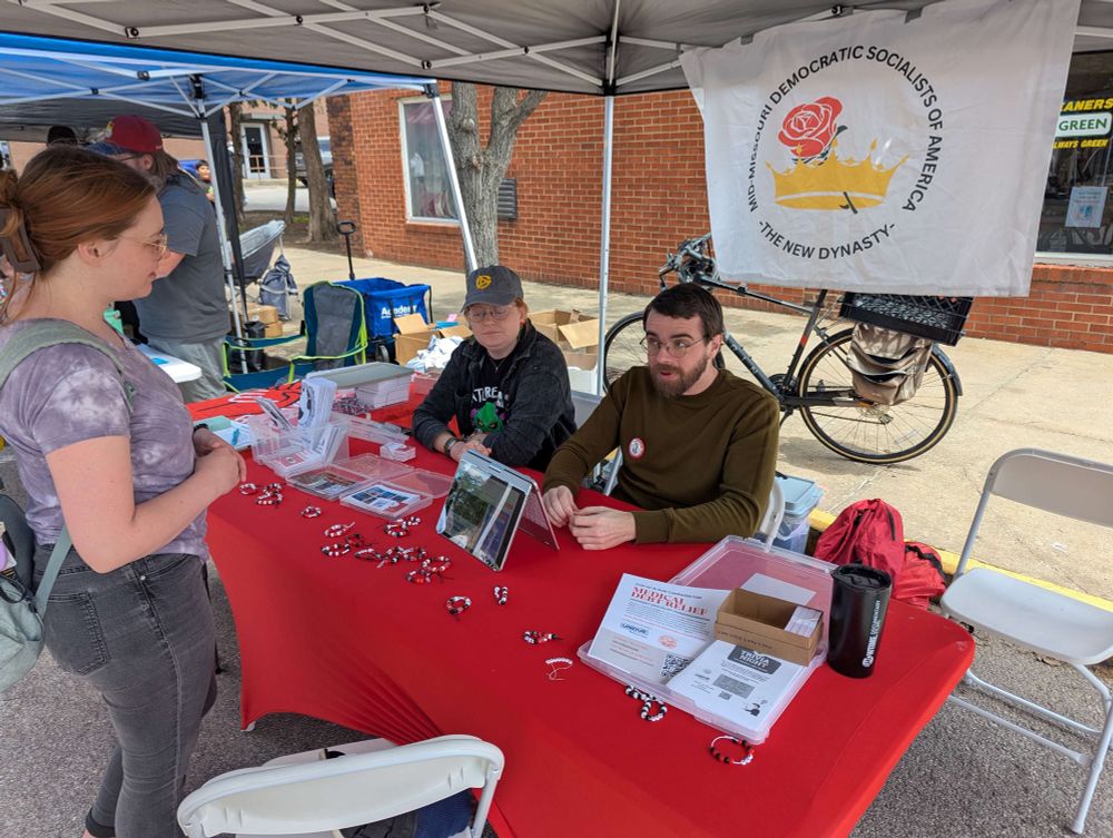 Three people sit behind a red table under a canopy at a Mid-Missouri Democratic Socialists of America (DSA) booth, engaging with a visitor standing in front of the table. The table is covered with pamphlets, buttons, and display materials. A DSA banner hangs on the back of the tent, and a bicycle is parked nearby.