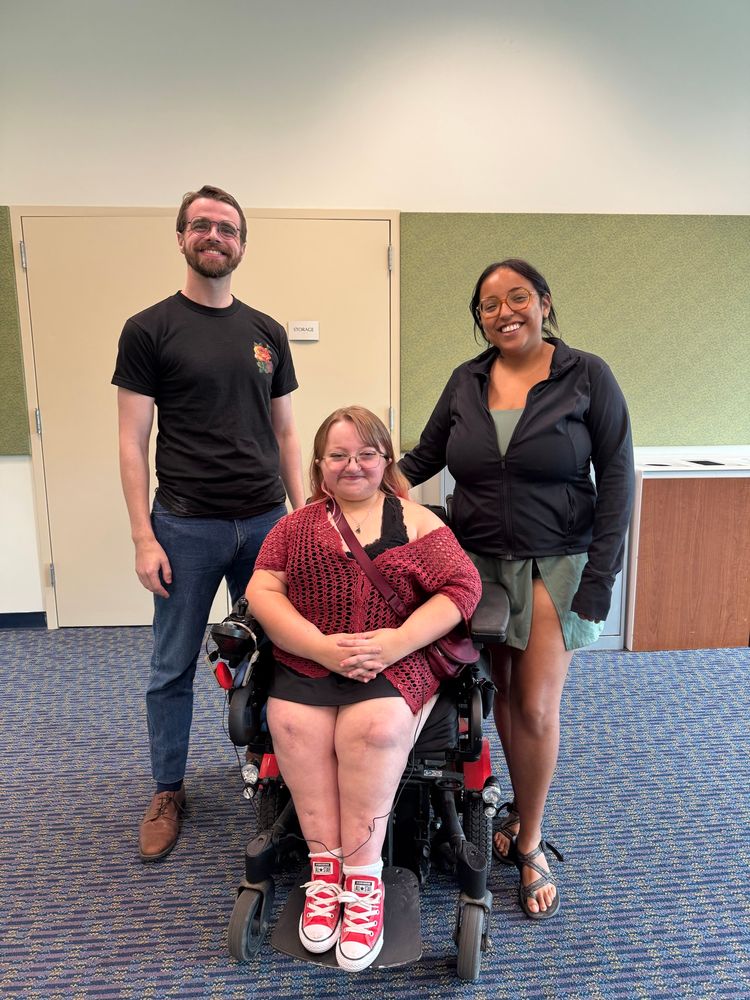 Adam, Sami, and Kenzie smile for a photo inside the library. Sami, a light-skinned woman in a power wheelchair, is between Adam, a light-skinned man in a black t-shirt, and Kenzie, a medium-brown-skinned woman in a black jacket and green skirt 