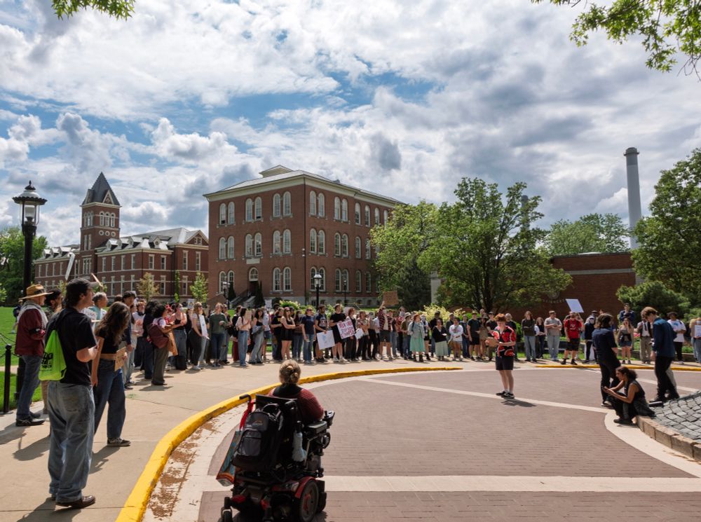 A large group of students stands in a semicircle outside a red-brick campus building during the MU Stand Up for Students walkout. Mizzou YDSA Chair Sammi, in a power wheelchair, faces the crowd while another speaker addresses the group. The sky is bright with scattered clouds, and the energy is focused and serious.