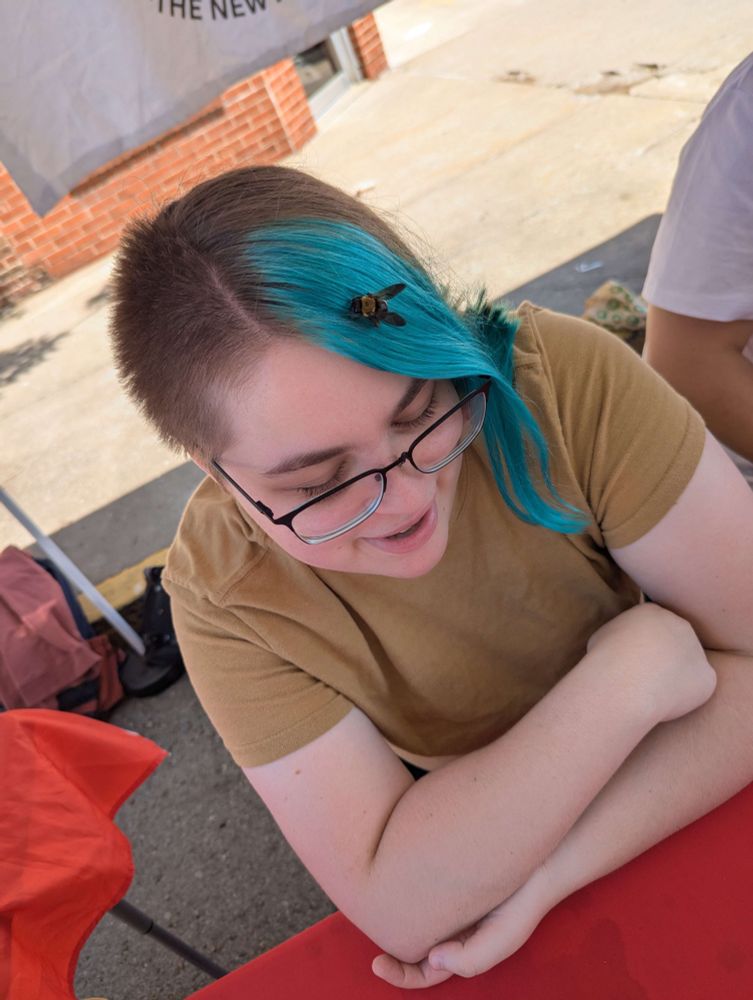 A young Person sitting at a red table outdoors at the Mid-Missouri DSA tent, smiling and looking down. They have short brown hair with one side dyed bright teal, and a bumblebee is resting on their hair. They are wearing glasses and a brown t-shirt.