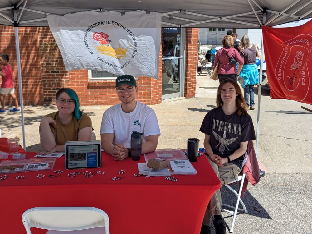 Three people sit behind a red table under a canopy at the Mid-Missouri Democratic Socialists of America (DSA) booth. The table is covered with buttons, flyers, a tablet, and other materials. Two DSA flags are displayed behind them, one white and one red. They are smiling and seated in front of a brick building as people walk by in the background.