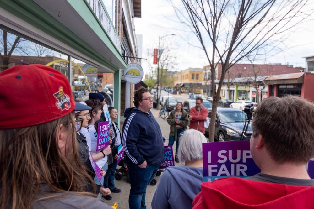 Alejandro, a restaurant worker, speaks to a diverse crowd outside Yellow Dog Bookshop in downtown Columbia, Missouri. Supporters hold signs reading “I SUPPORT PAID SICK DAYS” and “I SUPPORT FAIR WAGES” as media records the workers’ rights rally.