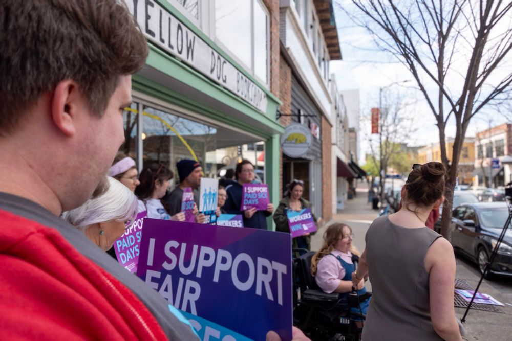 A diverse group of people stands outside Yellow Dog Bookshop in Columbia, Missouri, holding bright signs that read “I SUPPORT PAID SICK DAYS” and “WORKERS WIN.” A woman in a wheelchair is near the front, while others of varying ages and backgrounds line the sidewalk. A media tripod is set up to capture the gathering.