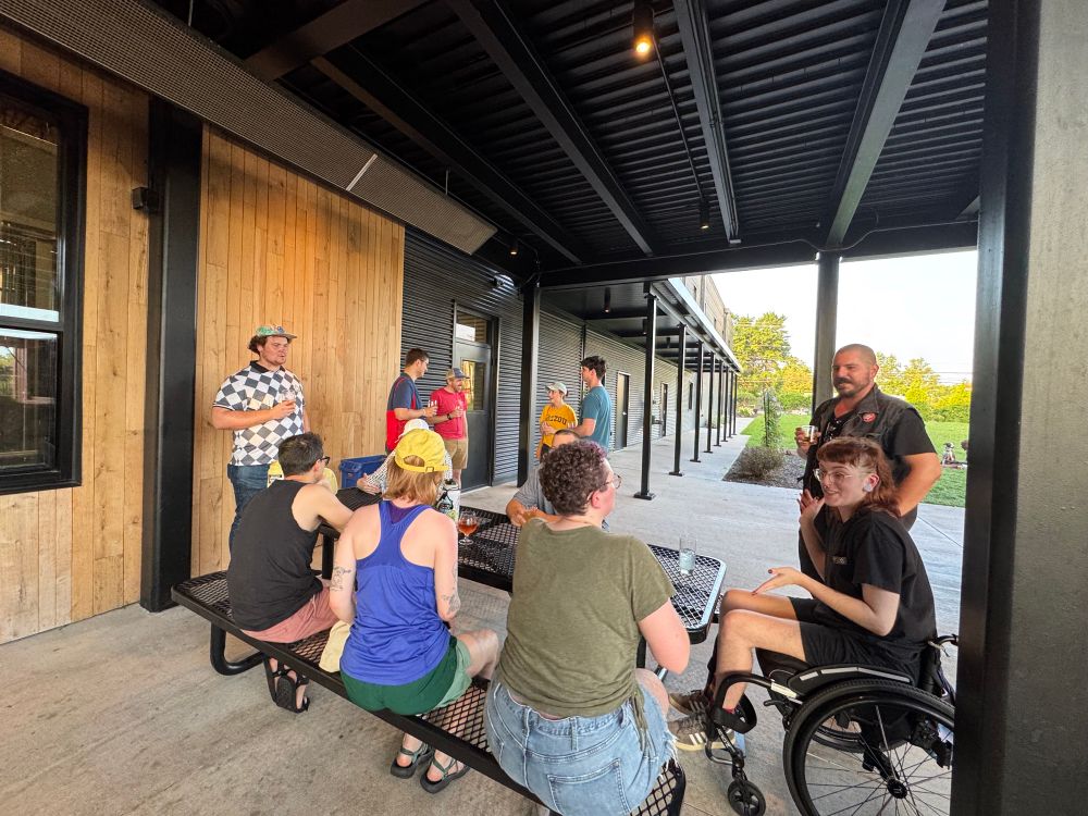 A group of people sitting at a table at Logboat. One person uses a wheelchair while six others are standing. 