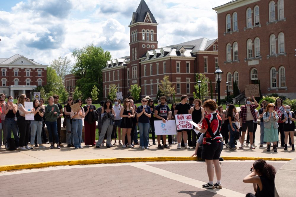A large group of students stands in a semicircle outside a red-brick campus building during the MU Stand Up for Students walkout. Mizzou YDSA Chair Sammi, in a power wheelchair, faces the crowd while another speaker addresses the group. The sky is bright with scattered clouds, and the energy is focused and serious.
