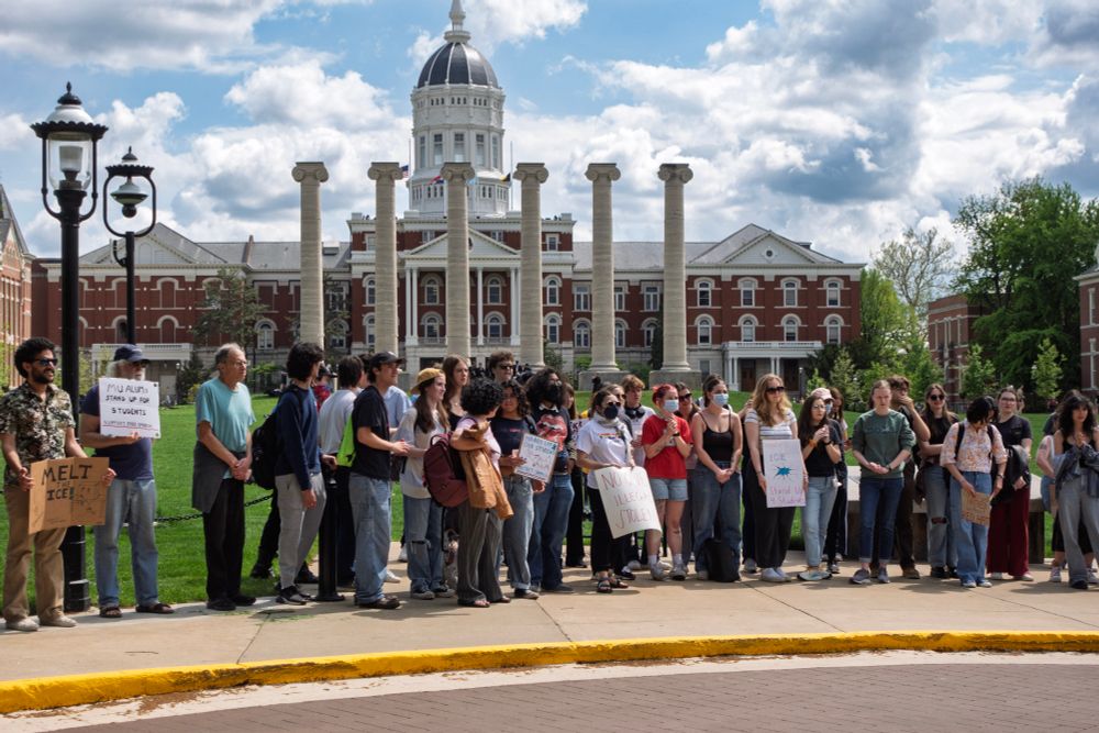 A large group of students stands shoulder to shoulder in front of the iconic columns and Jesse Hall on Mizzou’s campus during the Stand Up for Students walkout. Many hold protest signs, including one that reads “MU CAN’T SILENCE US” and another that says “MELT ICE.” The sky is partly cloudy, and the moment captures a strong sense of unity and resistance.