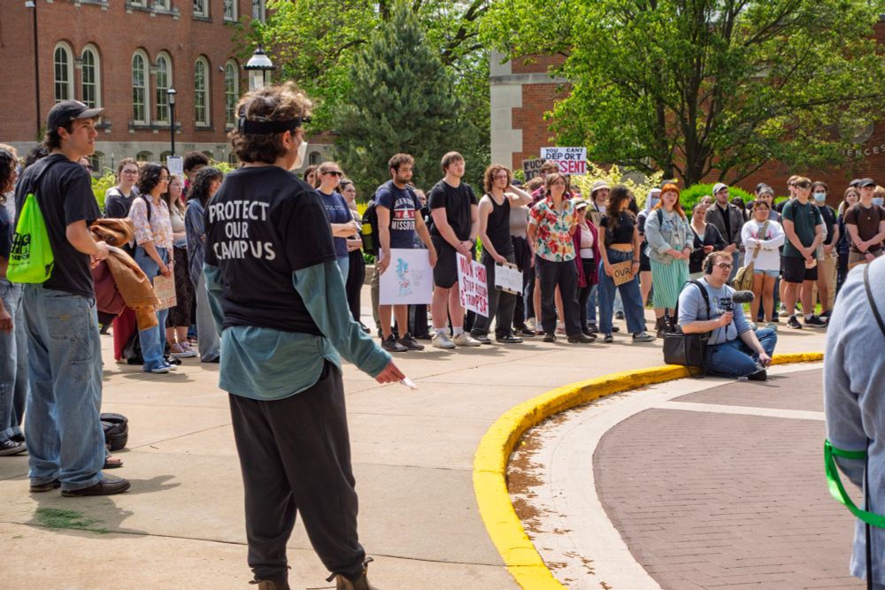 A group of students gather in a circle outside on a sunny day during the MU Stand Up for Students walkout. One student in the foreground wears a black shirt that says “PROTECT OUR CAMPUS.” Many participants hold signs and listen attentively, surrounded by trees and campus buildings.