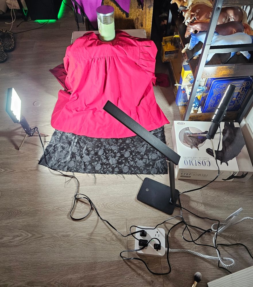 A photo of the setup I'm using for model horse photographs today. There is a red button down blouse draped over an empty box, held in place by a candle. A rectangle of black satin fabric with a rose pattern is underneath it, on the floor. One LED light is on the floor to the left. A second LED light is on the floor in the front, just to the right. A third LED light is on a box, on the right. You might notice the makeup brush near the bottom of the photo; this is for brushing dust off the models.