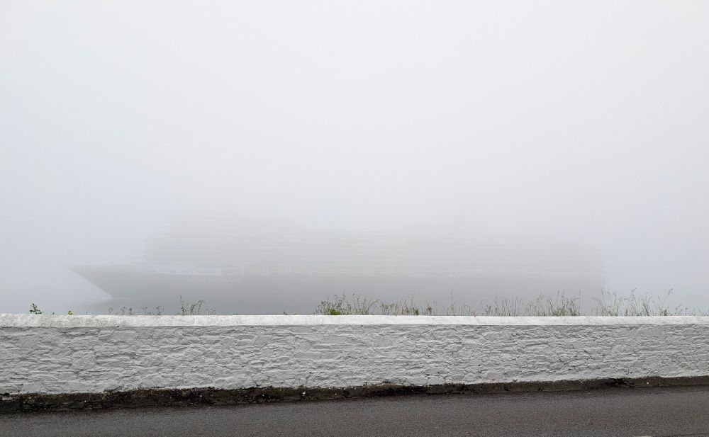 Very foggy view with a barely discernable outline of the Holland America Line cruise ship leaving Cobh. The foreground shows the road and white sea wall but very little is visible above the sea wall because of the fog. 
