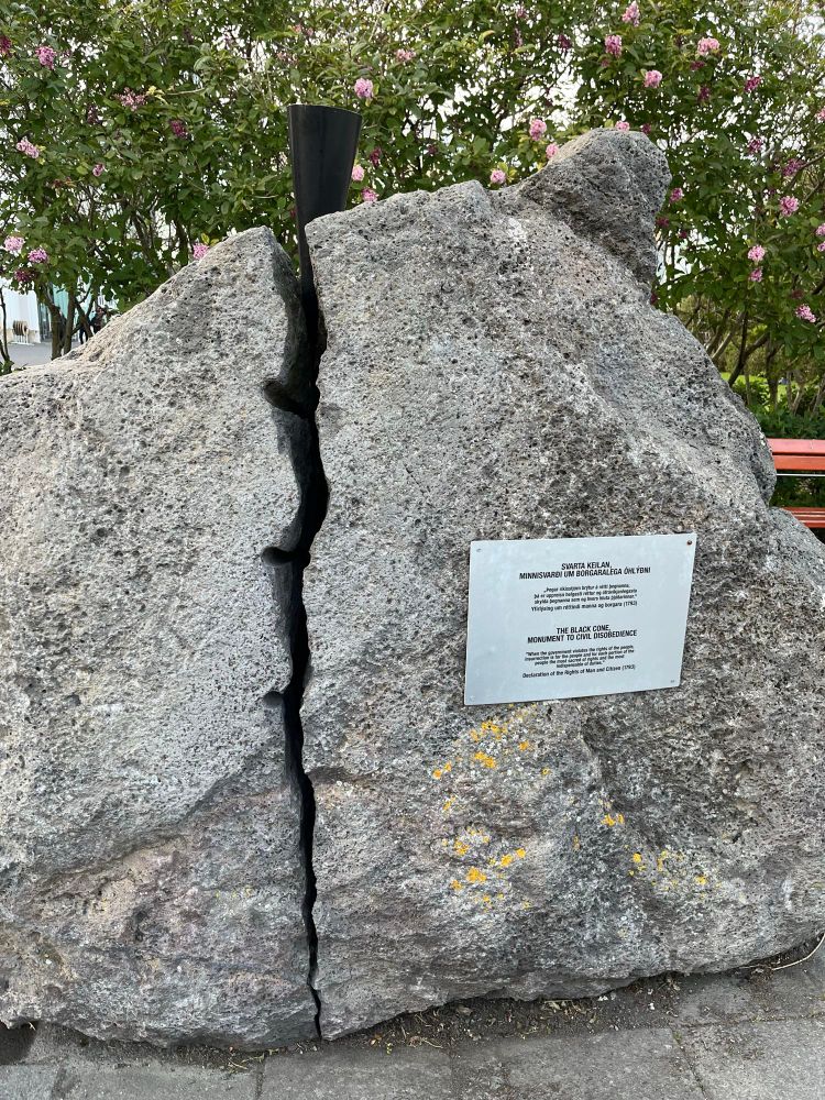 Photo of a large stone, splitting down the middle, a black cone is wedged in the crack. A plaque on the rock gives the title and inscription of the piece.