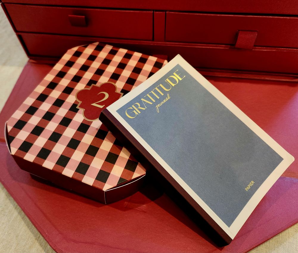 A small paperback journal with a blue cover rests against a red gingham-patterned box with the number 2 on it. "Gratitude" is written in yellow on the journal's cover