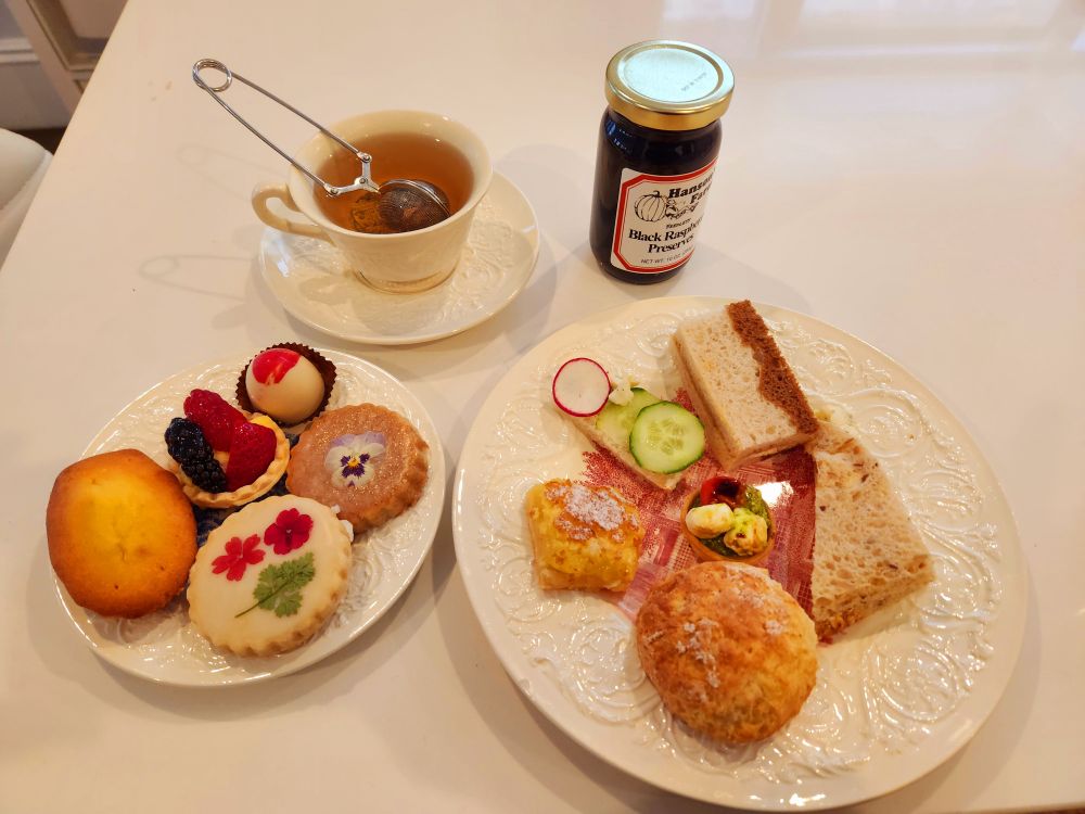 A cup of rose tea steeps next to plates of savory and sweet teatime delicacies and a jar of local farm-made blackberry preserves