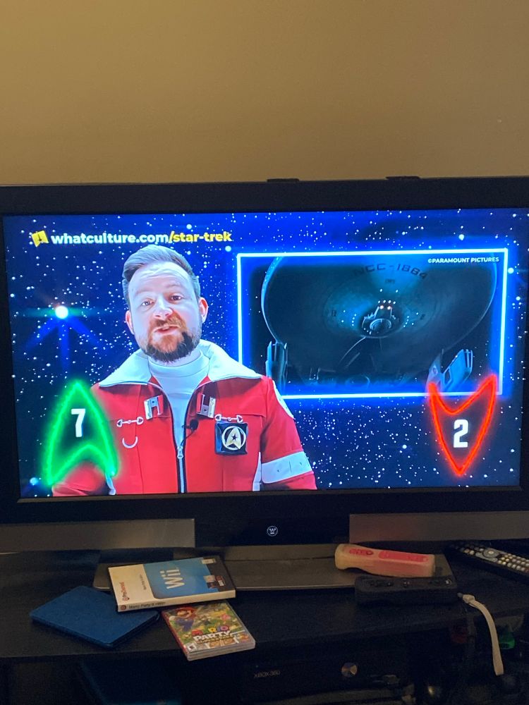A TV showing an episode of TrekCulture where Sean is wearing a Star Trek uniform from the movie Star Trek 2. Sub image shows a picture of the USS Reliant from this movie. There are 7 up Deltas and 2 down Deltas. Also around the TV is a WiiMote and Wii game, a Kindle, TV remote, and a Switch game “Mario Party Superstars”.