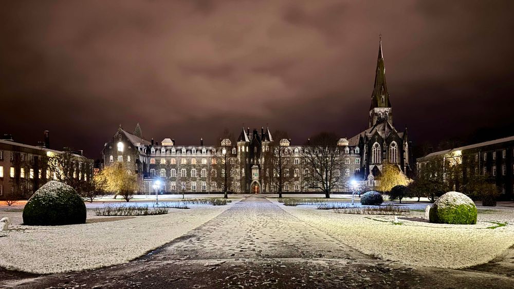 A night shot of a large quadrangle with a three-story revivalist gothic building on the opposite side with a church with spire to it’s left. The quad is covered in a layer of snow, with footprints clearly visible in the wide path running from the front of the frame through the middle of the quad to the opposite building. A clouded sky above glows orange with light pollution.