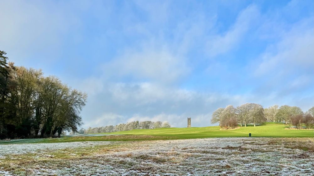 A wide landscape shot looking up a hill with a square crenellated tower on the horizon at the top of the slope. The foreground is frozen grass, the mid-ground is green sun-lit grass, and there are stands of threes in the left mid ground and on both sides of the tower. A blue sky with thin fluffy off-white clouds dominates the top half of the shot.