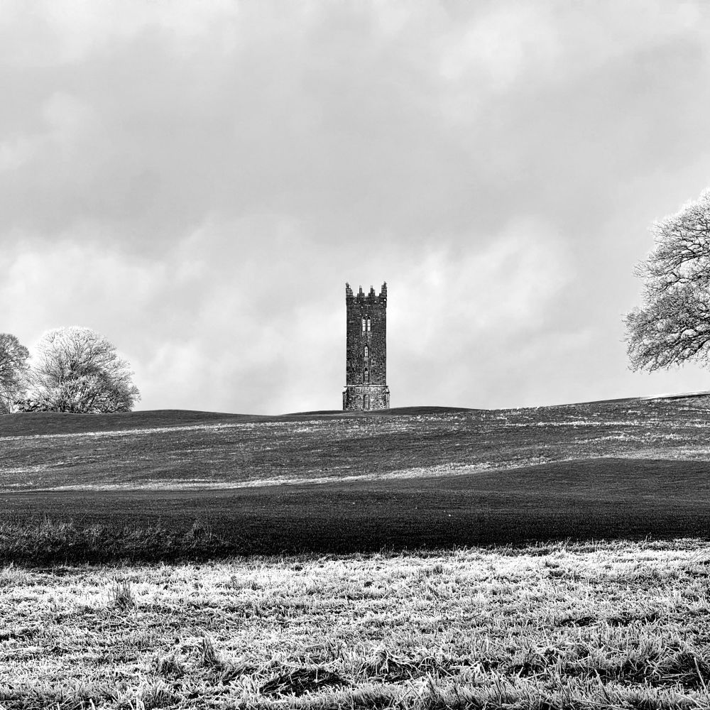 A square monochrome shot of a three-story square crenellated tower on the crest of a hill with a cloudy sky behind. There are frost-covered trees framing the left and right of the horizon, and white frosted grass in the foreground.