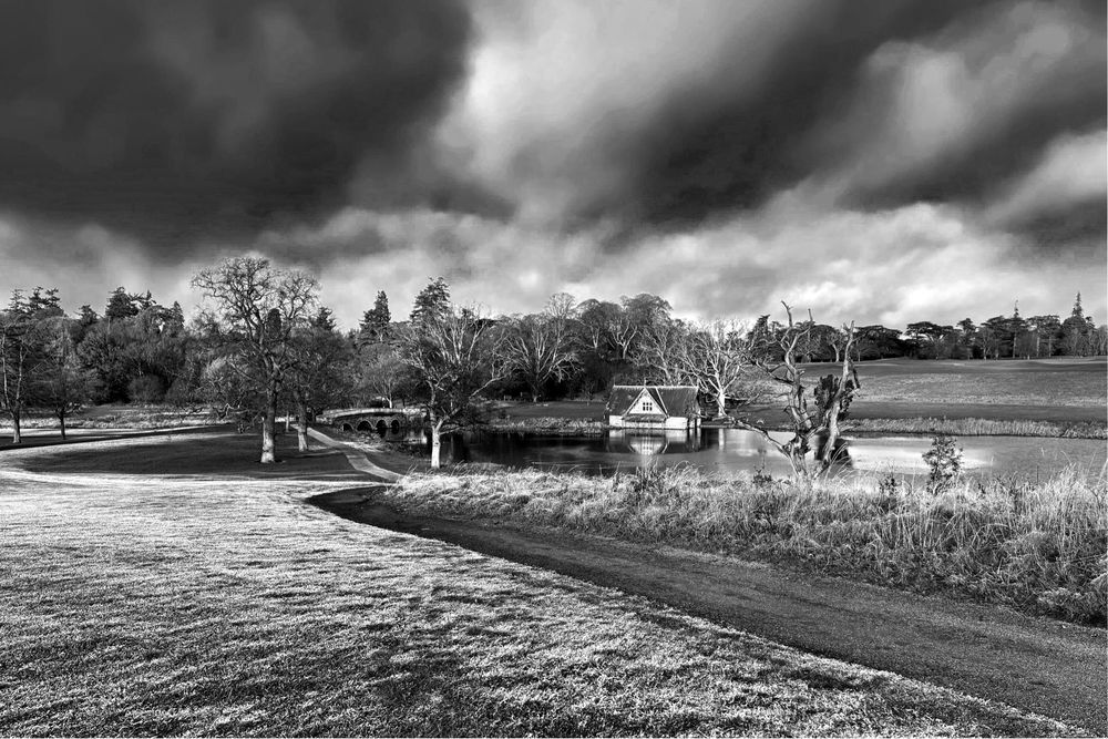 A black & white landscape shot looking down a slope with a winding path through frost-covered grass towards a small lake with a stone boathouse on the far bank. Behind the boathouse is another grassy bank and stands of trees. Overhead is a sky with white clouds against an inky black background.