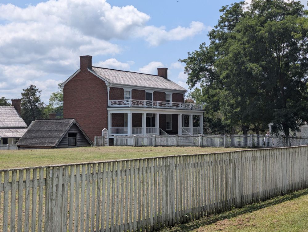 Brick house in the center of the photo. This is the McLean house, the building where Lee surrendered. There is a fence in the foreground and a big tree to the right of the brick house. A smaller building is visible in front of the house to the left of it as well.