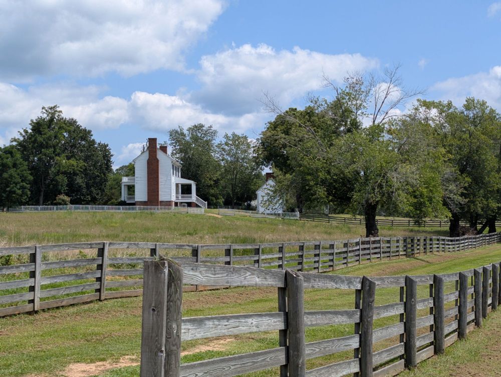 Grassy area with fences in the foreground. Closer to the background are trees, and prominent in the background is Isbell house. Mostly seeing the white walls and the brick chimney.