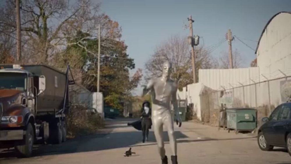 a man in a silver suit is running down a street next to a dumpster and a car .