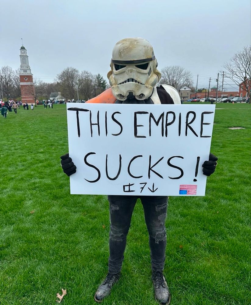 Person wearing stormtrooper armor with a sign that says "THIS EMPIRE SUCKS" 