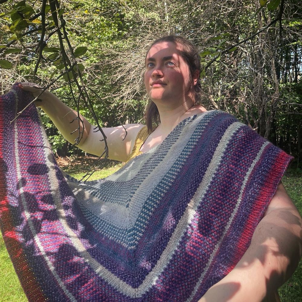 A white woman is holding a multi-colored hand-knit shawl under a tree; shadows of leaves and branches mark both the woman and the shawl. Her eyes look up to the sunlight. The shawl is striped with blues and purples with a hints of reds.