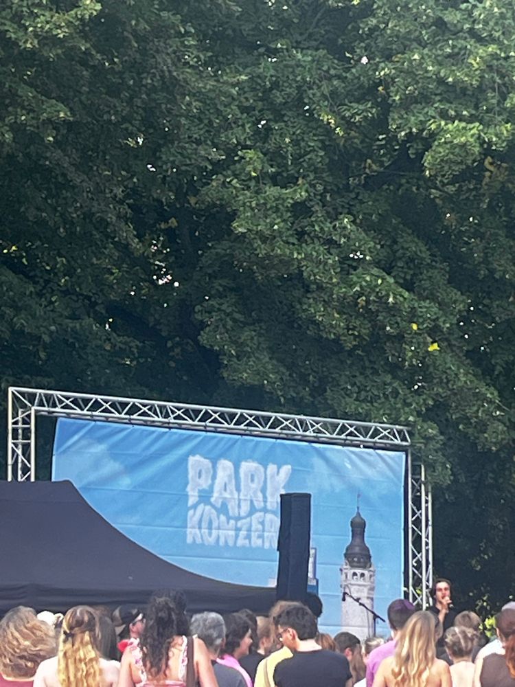 Bäume. Davor eine Bühne mit Banner als Rückwand, darauf steht „Parkkonzerte“ und ein Bild vom Neuen Rathaus Leipzig. Davor eine Menschenmenge.