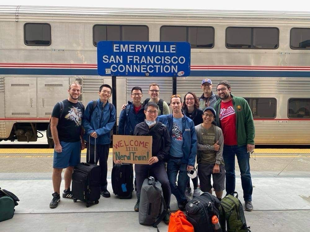 Photo of the nerdtrain group standing in front of an Amtrak train underneath the Emeryville San Francisco Connection sign. The group is surrounded by travel bags, and in the middle of the group is a person, Payton Chung, holding a sign that says Welcome Nerd Train!