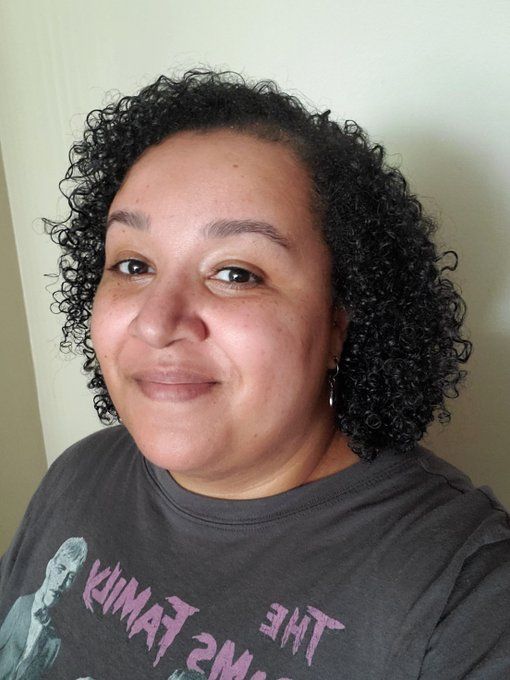 Danielle, a Black woman, in an adams family tee shirt smiling at the camera