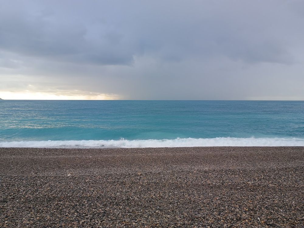 A rocky beach., a blue-green sea, a sky with some sun but a lot of clouds heavy with rain.