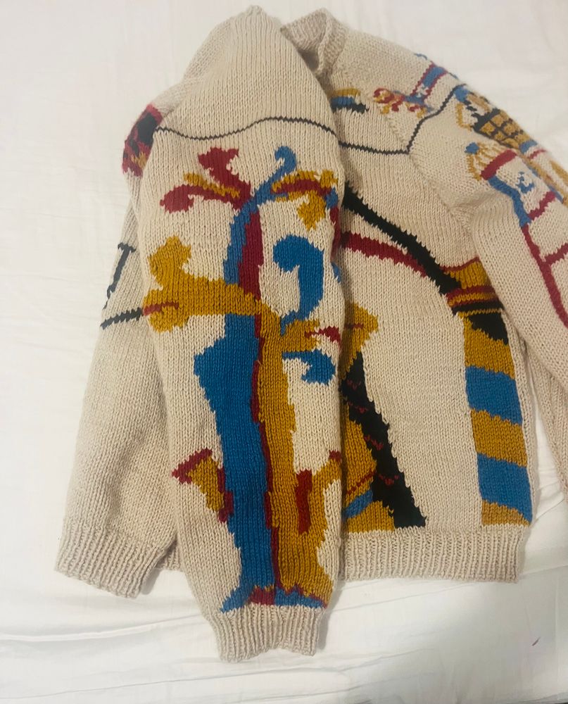 Beige jumper on a white background, a tree motif on a sleeve in blue, gold and red 