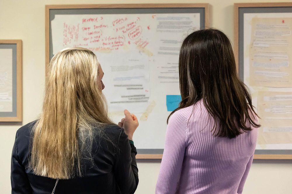 Two people with long hair have their backs to us. They are looking at a collage which is in a large wooden frame and hung on a wall.