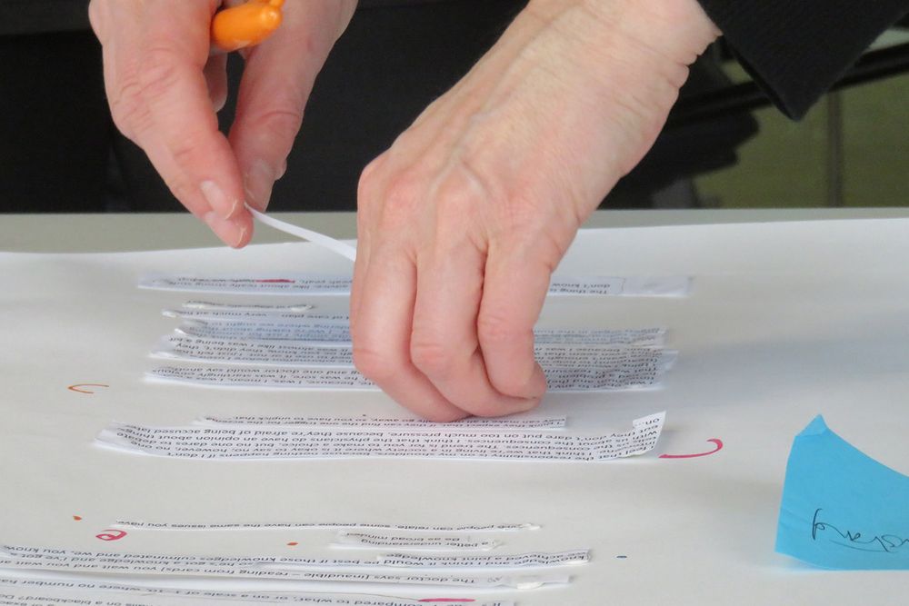 A pair of hands is sticking blocks of text to a larger sheet. One of the hands is holding an orange pen.