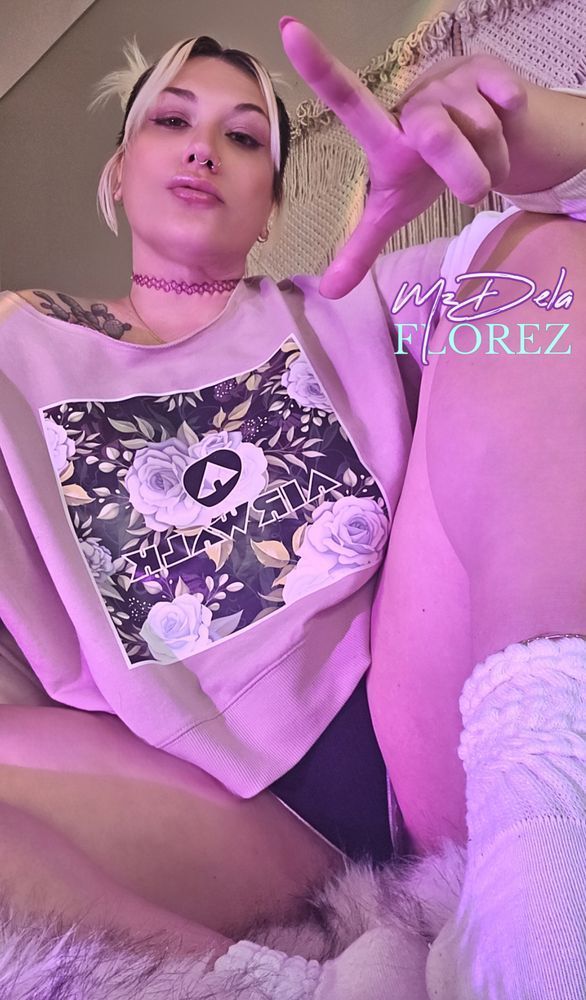 findom & femdom MILF Goddess, Mz Dela Florez, is dressed in a retro pink Y2K Airwalk off-the-shoulder sweatshirt, black short shorts with white athletic trim, and crisp white cotton slouch socks. Her platinum blonde hair is up in space buns, and She wears a pink jelly choker, as She makes commanding eye contact while making the L loser-sign with Her hand.