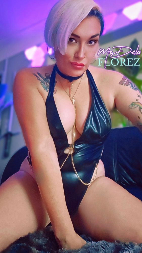 findom & femdom MILF Goddess, Mz Dela Florez kneels on a black leather couch, wearing a shiny black PVC bodysuit with a gold chain accent hanging between Her luscious natural tits. She makes intense eye-contact with a disappointed expression on Her face. you feel so small and somehow also aroused...