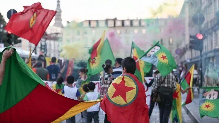 Kurdische Demonstrant*innen eingehüllt in Fahnen der PKK, im Hintergrund gelber, roter und grüner Rauch.