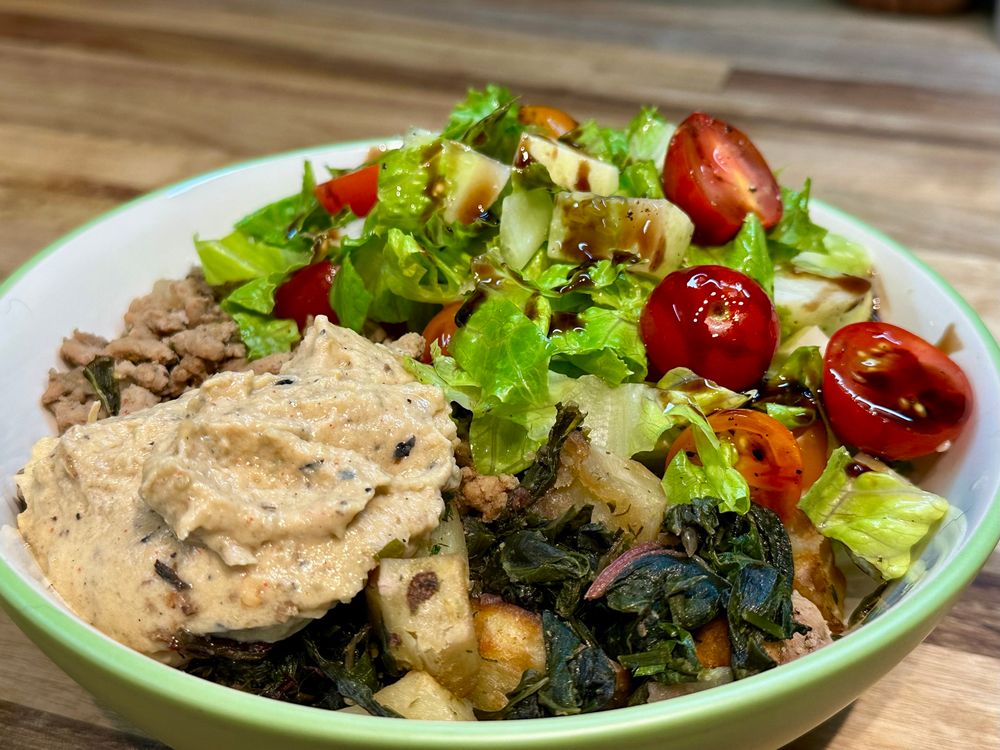 A green bowl half full of lettuce cucumber and cherry tomatoes, with a scoop of baba ganoush and cubes of potatoes, sautéed greens and ground turkey. 