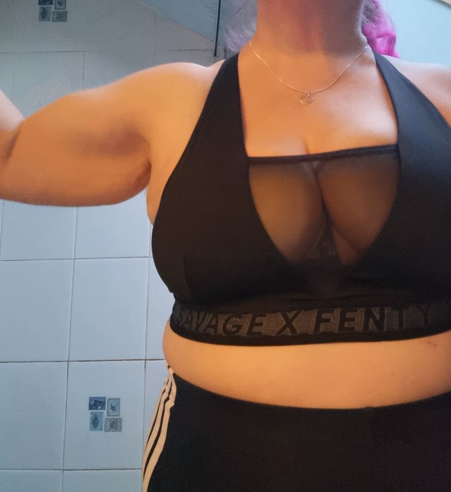 A woman in a sports bra and shorts is off centre in the image. She is stepped to the side to have her shoulder and arm in shot. The muscles in her shoulder and bicep are visible. She is strong.