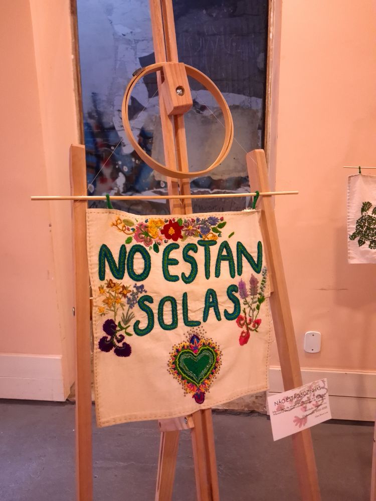 A imagem mostra os dizeres em espanhol "No están solas" (Não estão sozinhas, em português) bordados em um pano bege. Nele também está bordado um coração verde, flores amarelas, lilazes, vermelhas e rosas.