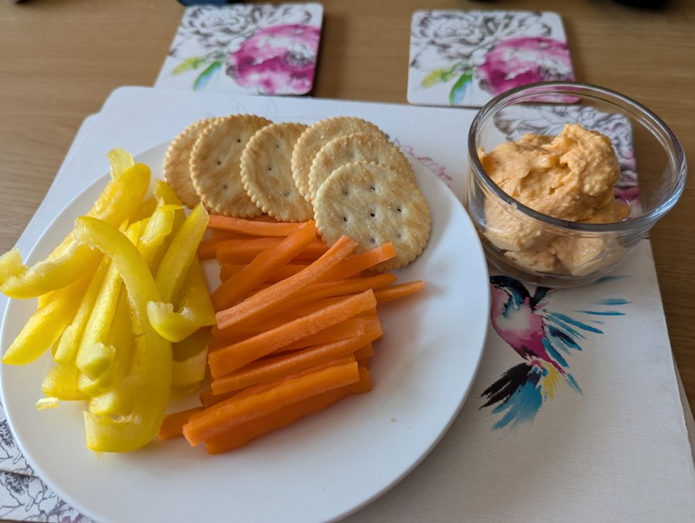 Carrot sticks, pepper sticks, and Ritz crackers on a small plate next to a pot of red pepper houmous.