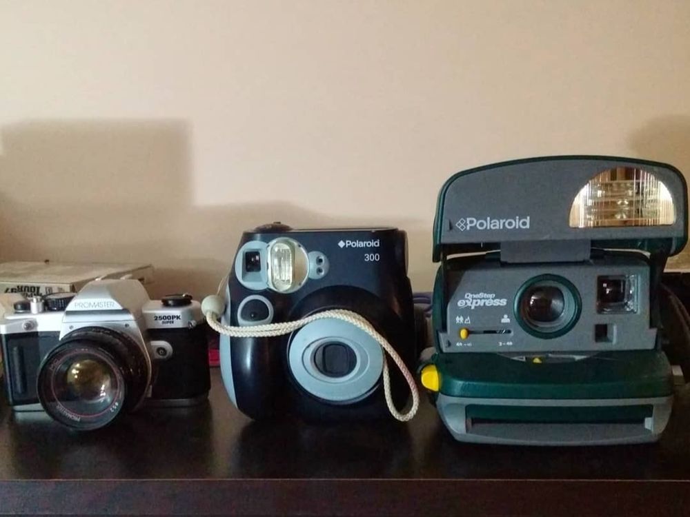Three cameras on a shelf. A 35mm film SLR film camera, an instax mini camera, and a Polaroid instant film camera 