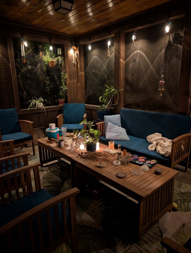 Screened porch at night, wooden furniture with teal cushions, plants, candles, and string lights. It's very cozy and a little cluttered.