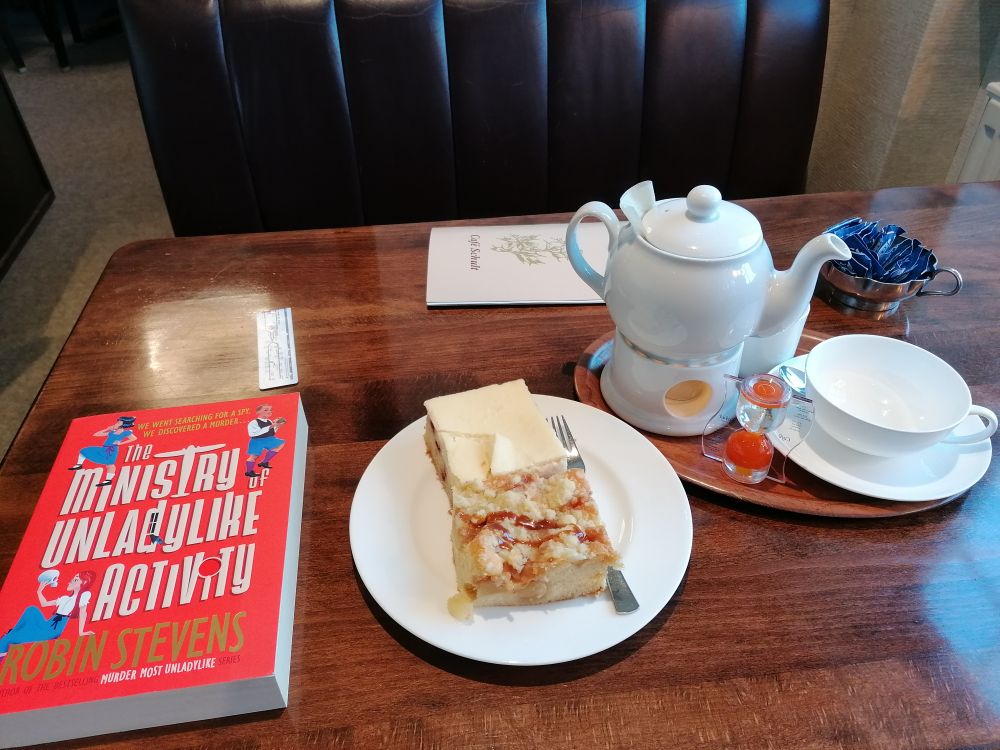 Cafétisch. Links Buch, mittig Teller mit Kuchen, rechts Teekanne und Tasse