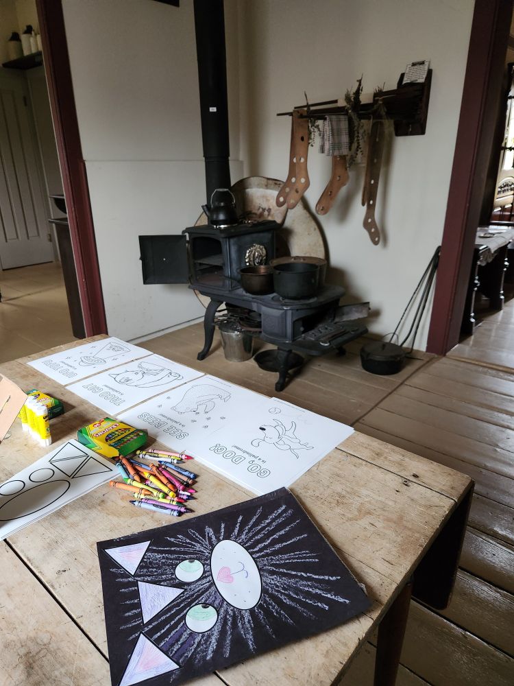 Cat-themed paper crafts, coloring sheets, and crayons are arranged on an old wooden table in a 19th-century kitchen. An old-fashioned wood-burning stove is in the background with a drying rack open above it.