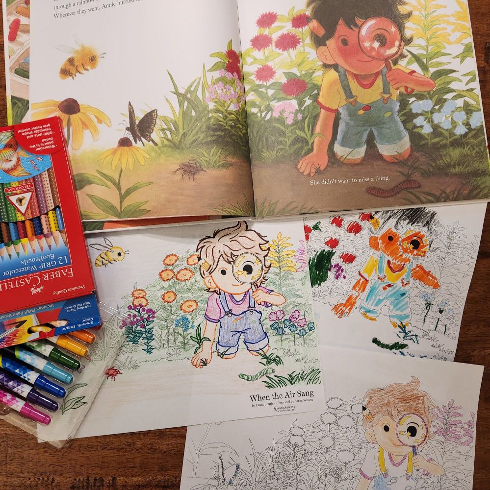 The picture book When the Air Sang is open to a spread showing a young girl kneeling in a garden surrounded by flowers and insects, looking at the reader through a magnifying glass. Beside the picture book are some pencil crayons and markers, and three coloring sheets featuring the scene from the book. Each sheet has been colored by someone of a different age with a different coloring style.
