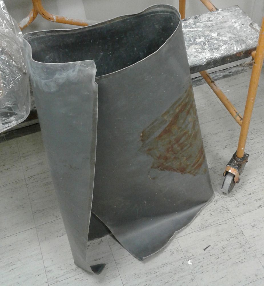 A large sheet of dull gray metal with a brownish splotch leaning up against a wheeled cart. It's about two feet tall and six feet long, and rolled up.