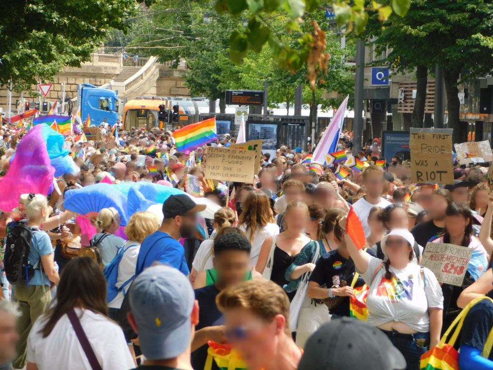 Zu sehen ist eine große Gruppe an Menschen auf der Demo mit einigen Prideflaggen, darunter unter anderem links mehrere Bi-Flaggen. Auch halten einige Menschen kleinere Plakate, auf denen unter anderem steht: "Feminism is my second favorite F-Word - first one is Familienrecht", "The first Pride was a Riot" und "Keine Kompromisse bei Menschenrechten - weg mit §218"